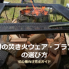 焚き火台の上で薪が燃え、オレンジ色の炎が上がっている様子。手前には割った薪が積まれ、周囲は芝生のキャンプサイト。難燃素材の焚き火ウェア・ブランケットの選び方というタイトルが写真下部に重ねられている