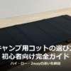 室内のフローリングに設置された、黒いキャンプ用コット(折りたたみベッド)の写真。キャンプ用コットの選び方 初心者向け完全ガイド ハイ・ロー・2wayの違いも解説というタイトルが写真下部に重ねられている